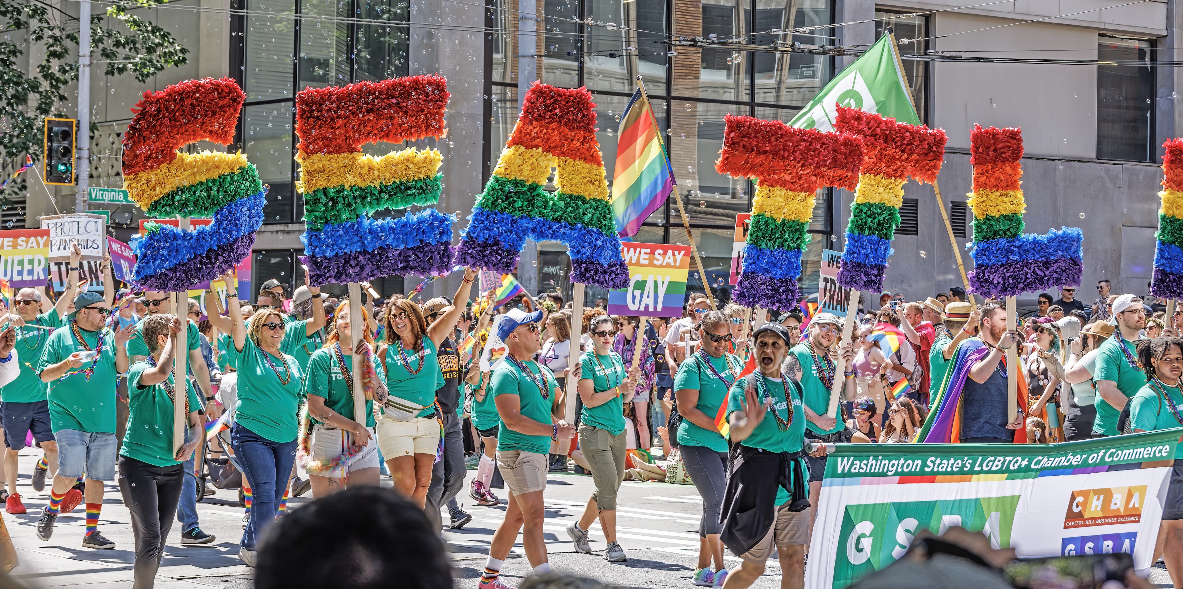 Photo of a pride parade at Mirabella Seattle. Participants are wearing teal t-shirts and holding up rainbow decorative letters that read Seattle.